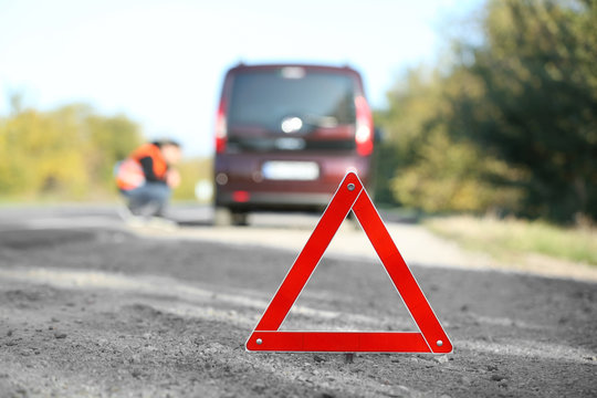 Red warning triangle on asphalt road. Tow truck worker near broken down car