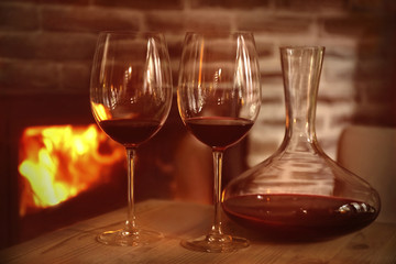 Decanter and glasses with red wine on wooden table against fireplace