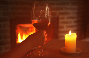 Female hand holding glass of red wine with blurred fireplace on background