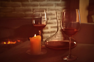 Decanter, glasses with red wine and candle on wooden table against fireplace