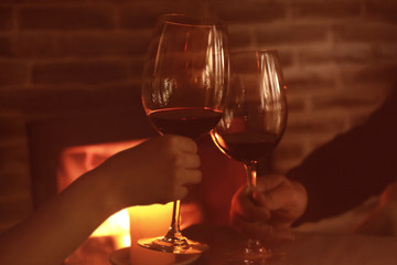 Hands of couple clinking glasses with red wine against blurred fireplace