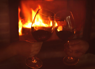 Hands of couple clinking glasses with red wine against blurred fireplace