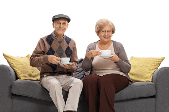 Joyful seniors with cups sitting on a sofa and looking at the camera