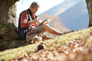 Hiker relaxing by tree looking at map and using tablet