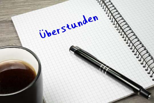 Ein Heft mit dem Wort &Uuml;berstunden und eine Tasse Kaffee auf einem B&uuml;rotisch