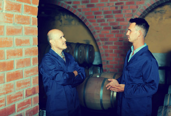 Fototapeta na wymiar two smiling men in uniforms standing in cellar with wine woods
