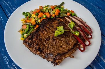 Grilled steak with steamed vegetables on a white plate. blue table. Top view