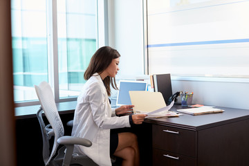 Female Doctor Wearing White Coat Reading Notes In Office