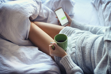 young woman drinking coffee in her bed and checking her smartphone