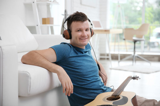 Young man sitting with a guitar on the floor and listening to music on headphones