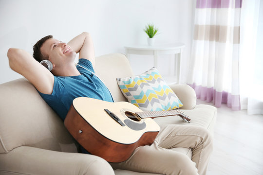 Young man with headphones playing guitar and sitting on a couch in light room