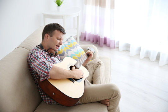 Young man with headphones playing guitar and sitting on a couch in light room