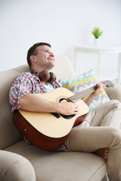 Young man with headphones playing guitar and sitting on a couch in light room