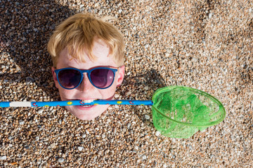 Boy with net in mouth buried in pebbles on beach