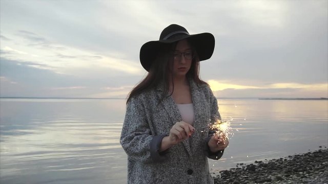 Pretty happy woman lights sparklers on the sea coast at sunset slow motion