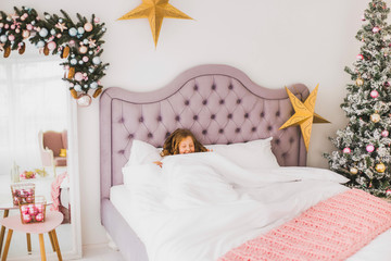 Portrait of cute funny little girl on Christmas morning in white home interior. Child pretending to be sleeping than rises cheerfully in bed. Horizontal color photo.