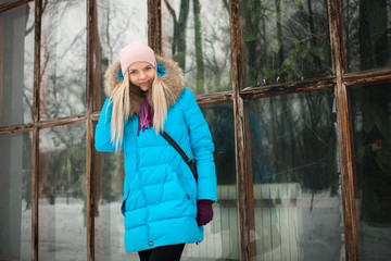 Fototapeta premium Cloudy outdoor winter portrait of young happy adorable woman in bright cyan coat posing in winter city park against large greenhouse window. Cold season lifestyle concept.