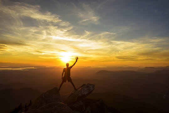 Silhouette hiker holding cap with hand raised while standing on top of mountain during sunset