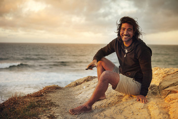 Portrait of happy man holding mobile phone while sitting on cliff by sea