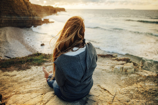 Rear View Of Woman Sitting On Cliff And Looking At View During Sunset