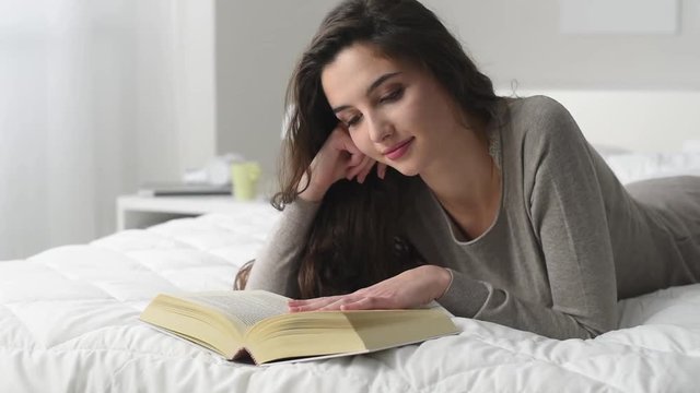 Young beautiful woman lying on the bed and relaxing, she is reading a book and smiling