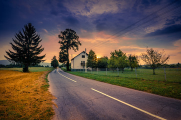 Road beside wheat field and cottage at sunset with colorful overcast sky.