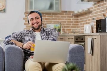 Portrait of happy laughing young man, student or freelancer businessman, works on his laptop and juice in hand at cozy modern home in loft style