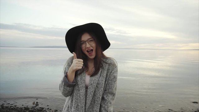 Young pretty woman in black hat and glasses showing thumb up near the sea at sunset