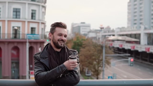 A stylish handsome man with a beard and trendy haircut smiling cheerfully, holding takeaway coffee or tea. Slow mo