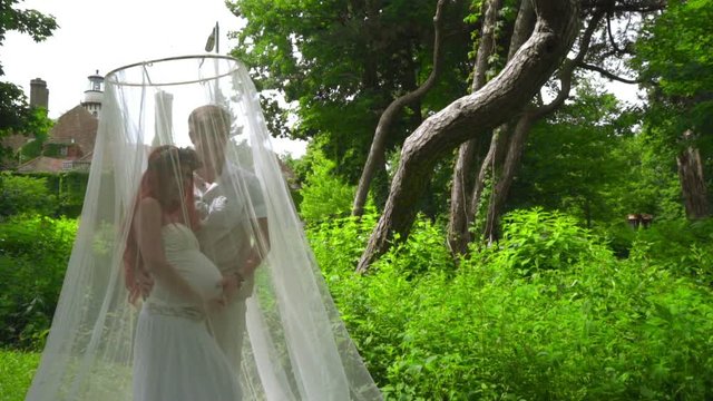 Pregnant couple posing under under wedding canopy at park. Couple in love in wedding decoration on green background. Man hugging red hair woman behind white fabric decoration. Wedding photo session