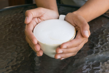 Beautiful asian woman holding hot cup of coffee with more milk foam on the glass table.