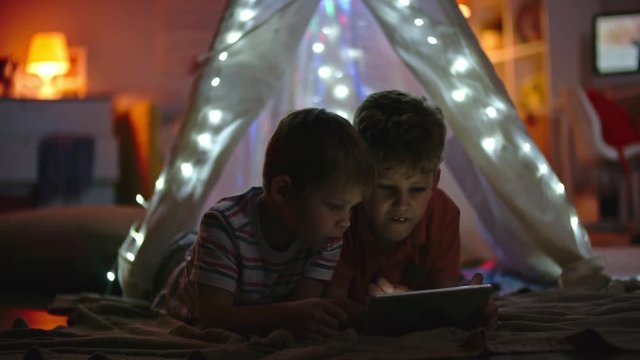 Little boys lying in teepee decorated with fairy lights and playing games on tablet in dark room