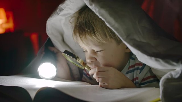 Little boy lying in bed under covers and reading book with flashlight and magnifying glass