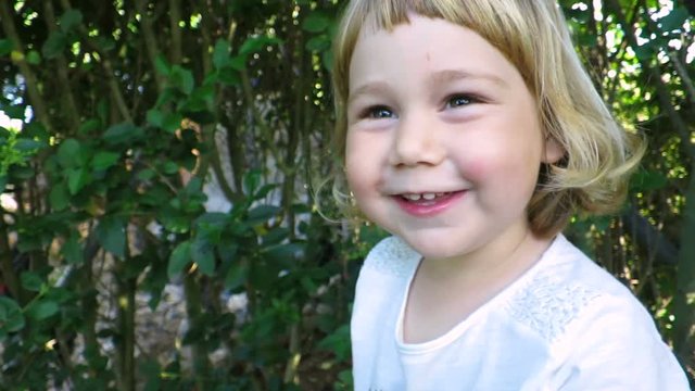 close up two years age blonde baby girl face talking, looking, teasing, sticking out tongue and hitting camera in green garden plants background
