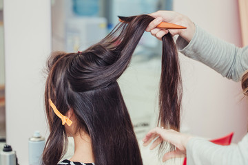 professional hairdresser doing a hairstyle to her client with healthy shiny brunette hair in the beauty salon, using comb and barrette