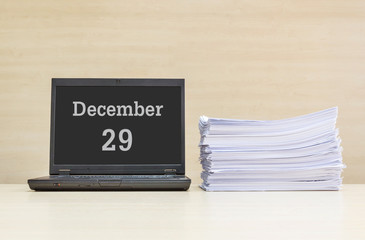 Closeup computer laptop with december 29 word on the center of screen in calendar concept and pile of work paper on wood desk and wood wall in work room textured background with copy space