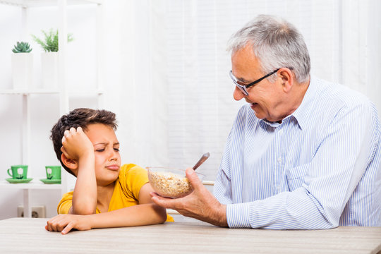 Grandfather is giving oatmeal to his grandson but he refuses to eat it. 