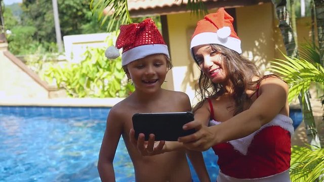 Happy smiling mother and child wear christmas hats taking picture self portrait on smartphone together in slow motion near the swimming pool on tropical island of Koh Samui. Thailand. 1920x1080