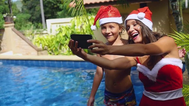 Christmas smiling mother and child wear christmas hats taking picture self portrait on smartphone together in slow motion near the swimming pool on tropical island of Koh Samui. Thailand. 1920x1080