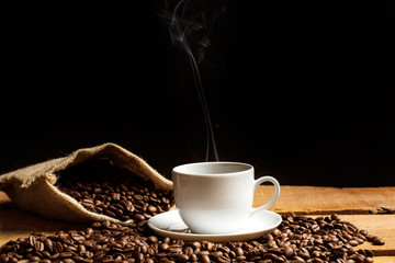 Taza de café con humo y granos de café con bolsa de arpillera sobre mesa de madera rústica y fondo negro. Vista de frente. Copy space