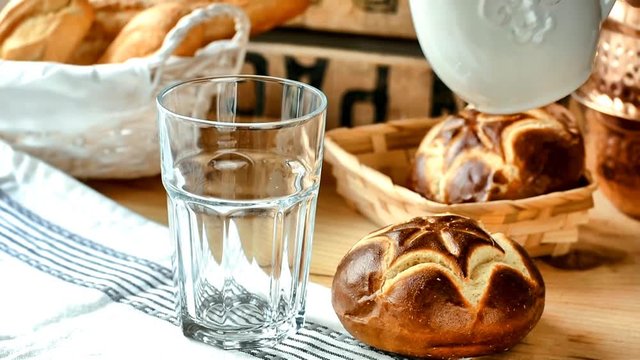 Process of pouring milk into a glass from pitcher,  fresh lye rolls in a wicker basket, wood table, rustic style kitchen interior, copper tableware in the background, cozy atmosphere, warm soft light