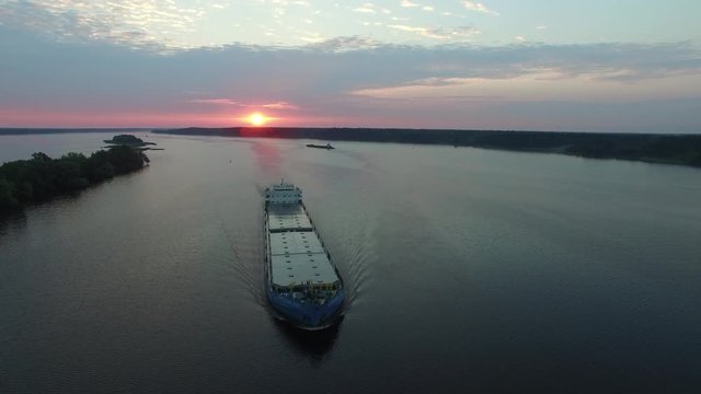 Cargo transport ship sailing on a wide river. Barge floating along the river at summer dawn. The camera moves in the air away from the ship. The island in the middle of the river. Aerial view.