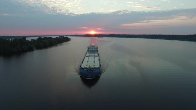 Cargo transport ship sailing on a wide river. Summer dawn. The camera moves through the air away from the ship. The island in the middle of the river. Aerial view.