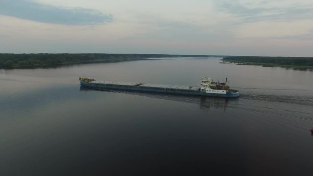 Cargo transport ship sailing on a wide river in the morningtime. The camera flying around the barge. Big transport vessel moving along the river. Aerial view.