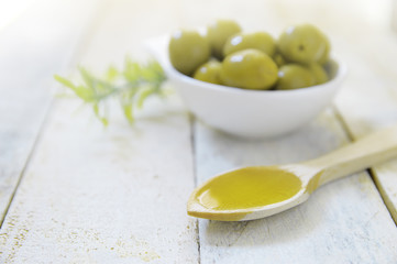 Spoon with olive oil next to some natural green olives. Scene on a wooden white table of a rustic kitchen.