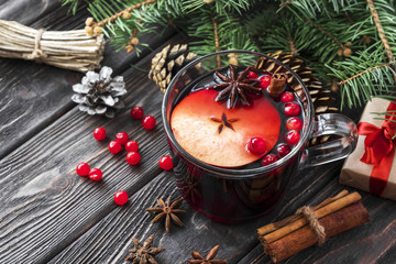 glass mug of mulled spicy red wine with red berries and apple on wooden brown table with cookies, cinnamon sticks and cones