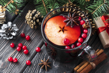 glass mug of mulled spicy red wine with red berries and apple on wooden brown table with cookies, cinnamon sticks and cones