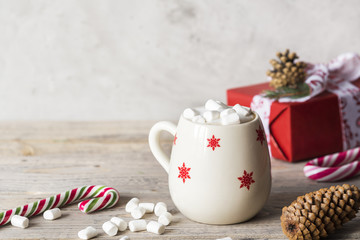 white cup with winter pattern with milky cappuccino on wooden light desk with christmas and new year decoration like candies, cones, berries and gifts wrapped in red paper