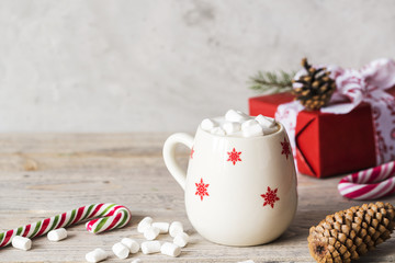 white cup with winter pattern with milky cappuccino on wooden light desk with christmas and new year decoration like candies, cones, berries and gifts wrapped in red paper