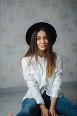 beautiful girl in a white shirt, blue jeans and a black hat sitting on the floor and looking into the camera. brunette model smiling from her bears ease